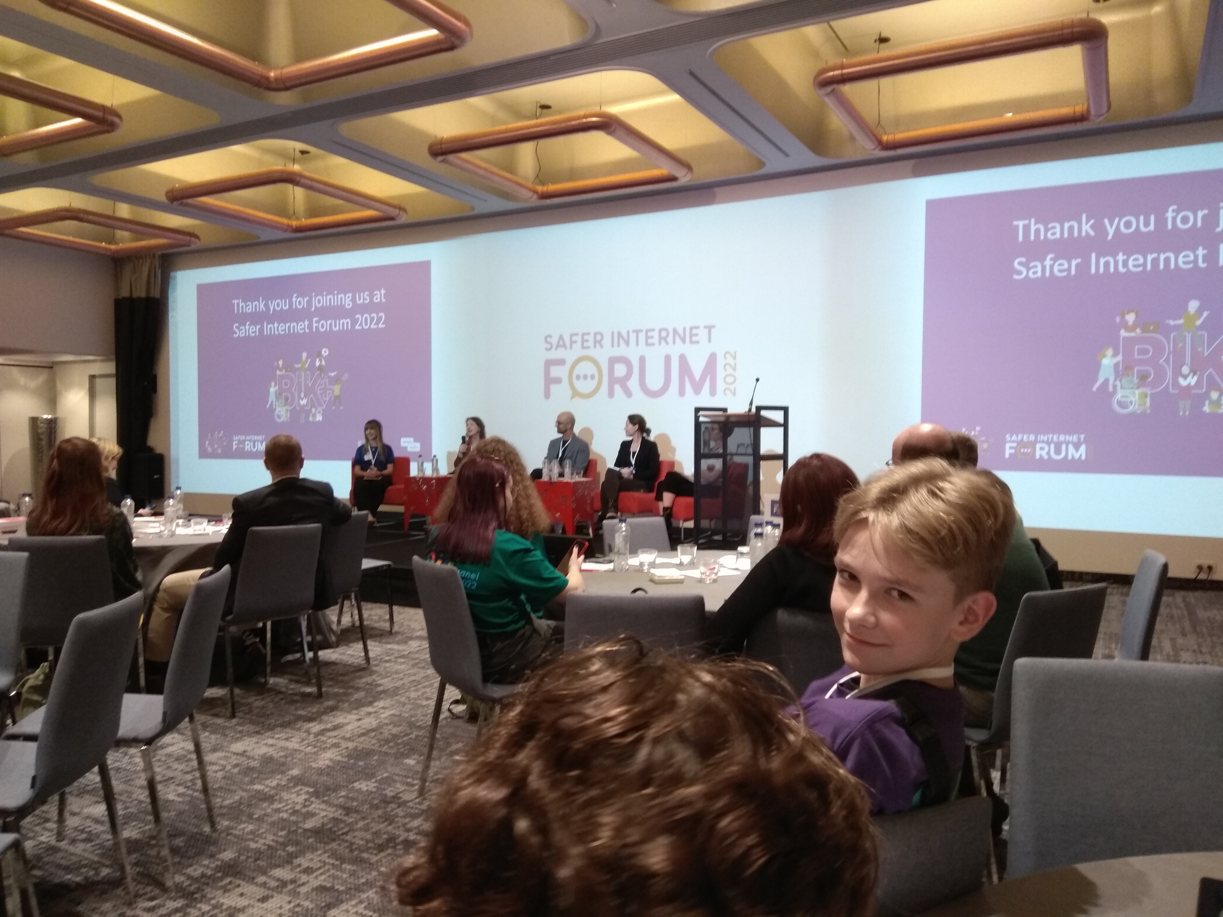 Sala konferencyjna, uśmiechający sie chłopiec, w&nbsp;tle wystąpienie prelegentów i&nbsp;napis Safer Internet Forum 2022